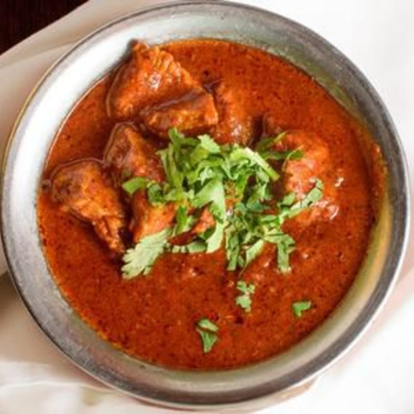 A metal bowl of rich red Indian curry with chicken pieces, garnished with fresh green cilantro.
