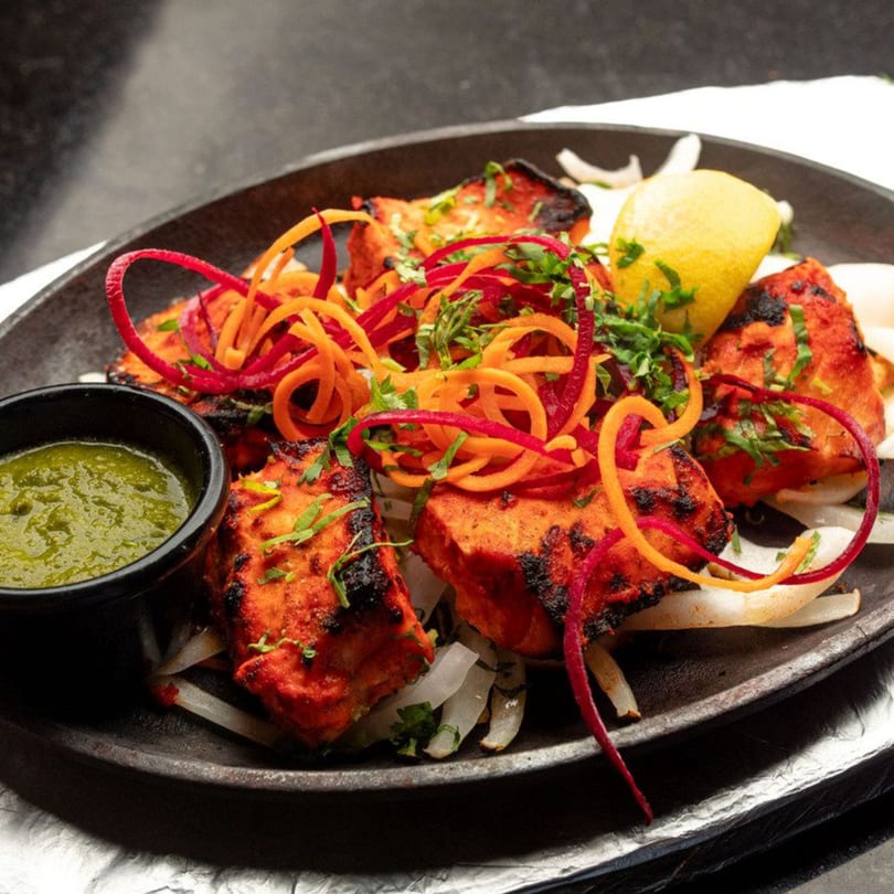 Indian chicken tikka with green chutney, lemon, and vegetable garnish on a dark plate.