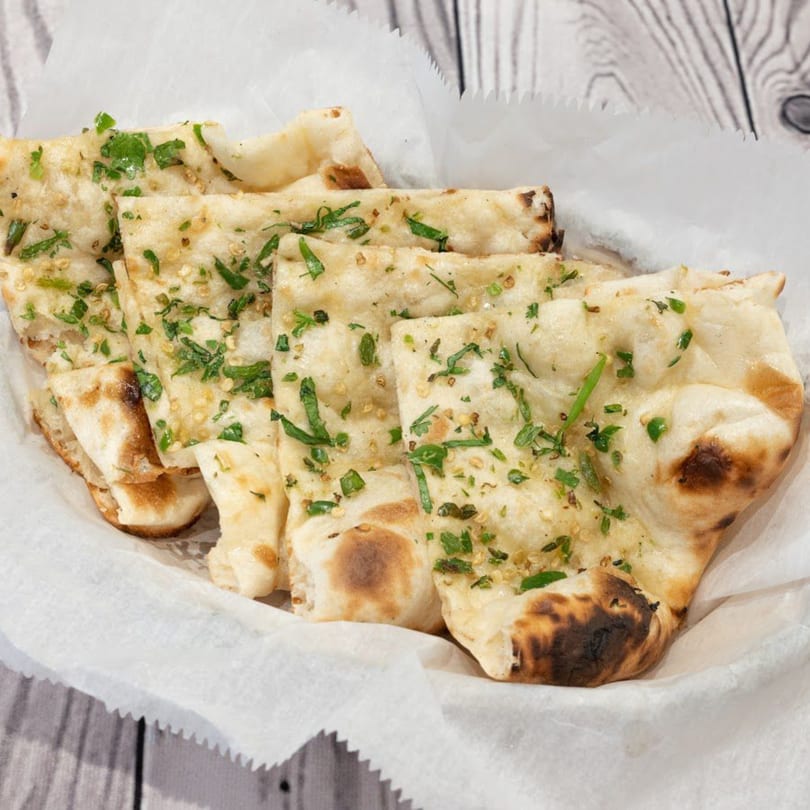 Indian garlic naan bread, garnished with green herbs and sesame seeds, served in a white paper-lined basket.